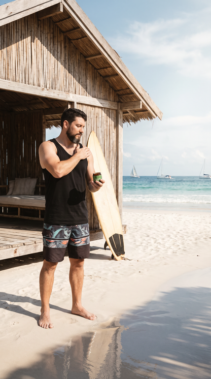 A man standing on a sandy beach in front of a wooden beach hut, holding a drink and a surfboard. He is wearing a black tank top and patterned swim trunks, with the ocean and sailboats in the background.