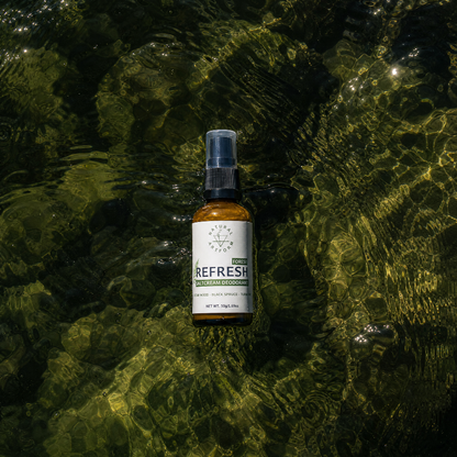 A small amber spray bottle labeled 'Pura REFRESH' and 'Forest Stream' floating on the surface of shallow, sunlit water with rippling reflections.