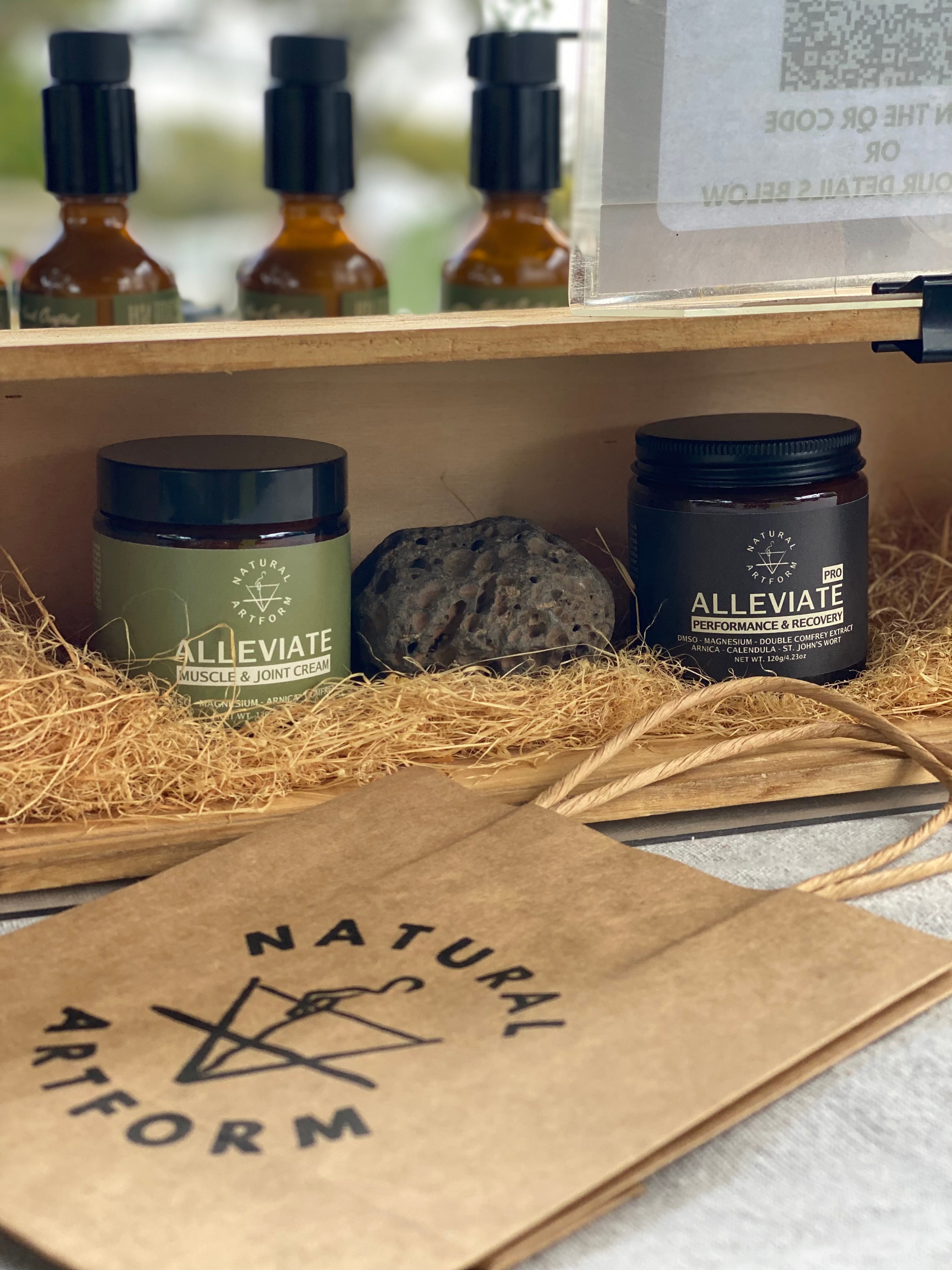 green label ALLEVIATE massage cream and ALLEVIATE PRO athletic body rub in hay straw with a recycled kraft paper bag showing Natural Artform alchemy logo